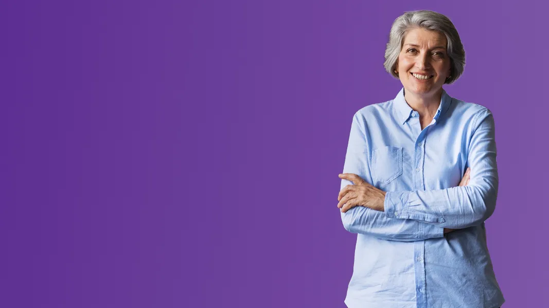 A woman in a smart shirt folds her arms and smiles in front of a purple background