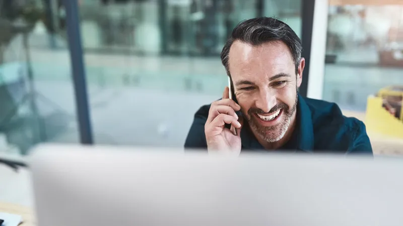 A man smiles while talking on his phone in front of a computer screen