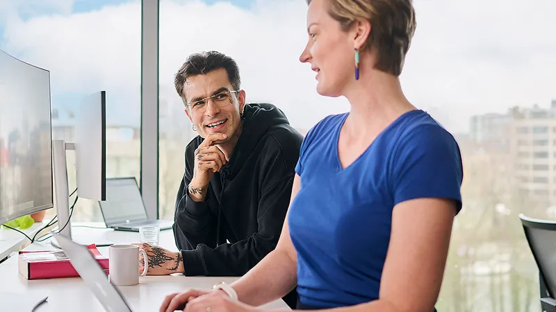 A man and woman are seated at a desk, engaged in a discussion while using a laptop.