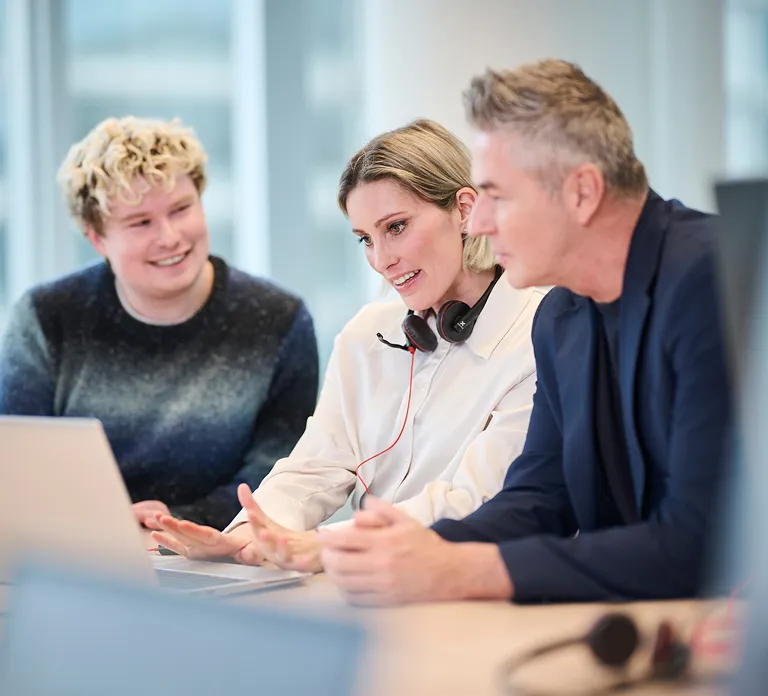 Three people collaborating at a table, focused on a laptop in front of them.