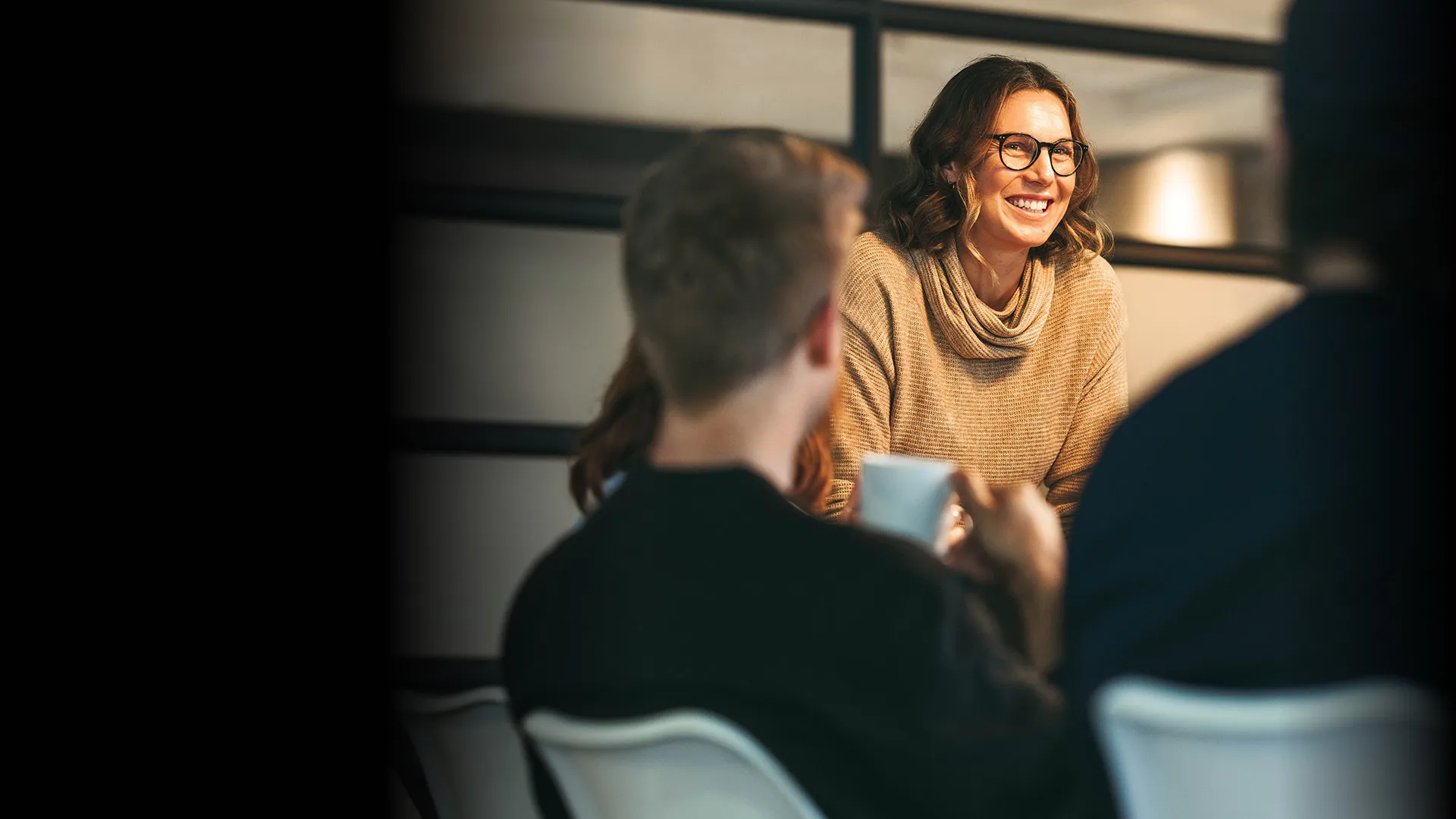 A woman smiles in a warmly lit office to colleagues across a table.