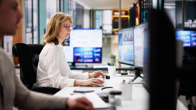 A woman looks at graphs on multiple screens while sat in an office setting