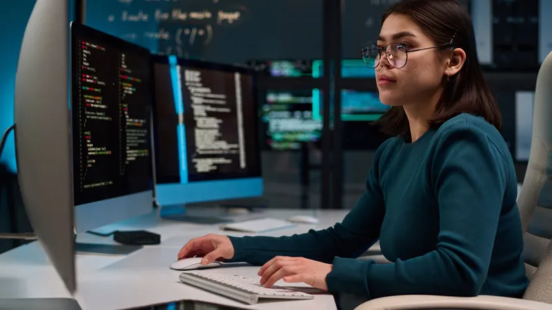 A woman looks at code on multiple screens in a dark office setting