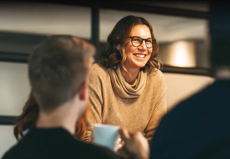 A woman smiles in a warmly lit office to colleagues across a table.