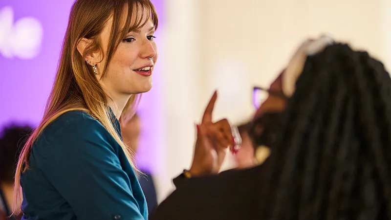 A woman converses with another woman at a social event, both engaged and smiling.